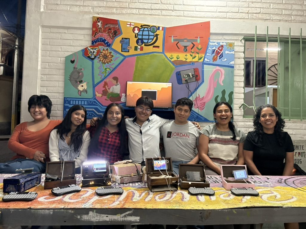 Siete personas sentadas en fila frente al tablero, sonriendo, con las computadoras artesanales abiertas sobre la mesa