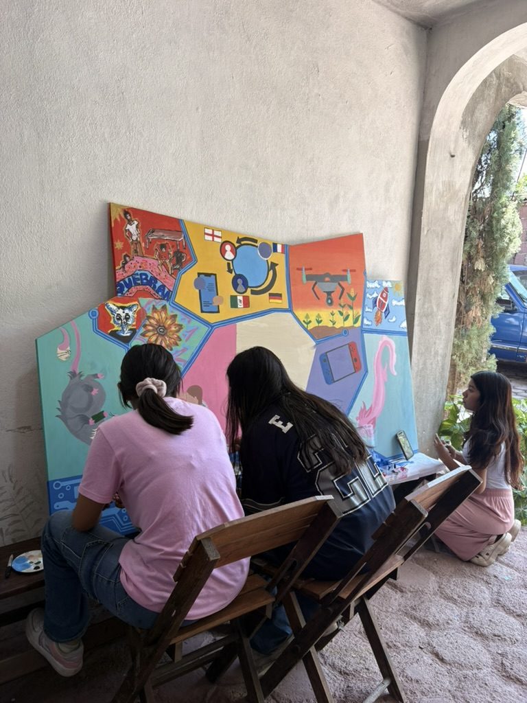 Dos jóvenes sentadas en sillas plegables pintan el tablero apoyado contra la pared; otra observa desde el suelo