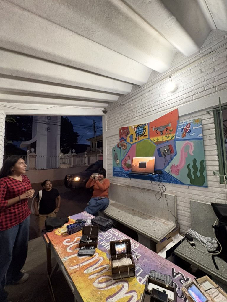 De noche, una joven explica de pie frente al tablero ya montado y encendido; sobre la mesa pintada, las computadoras en sus cajas