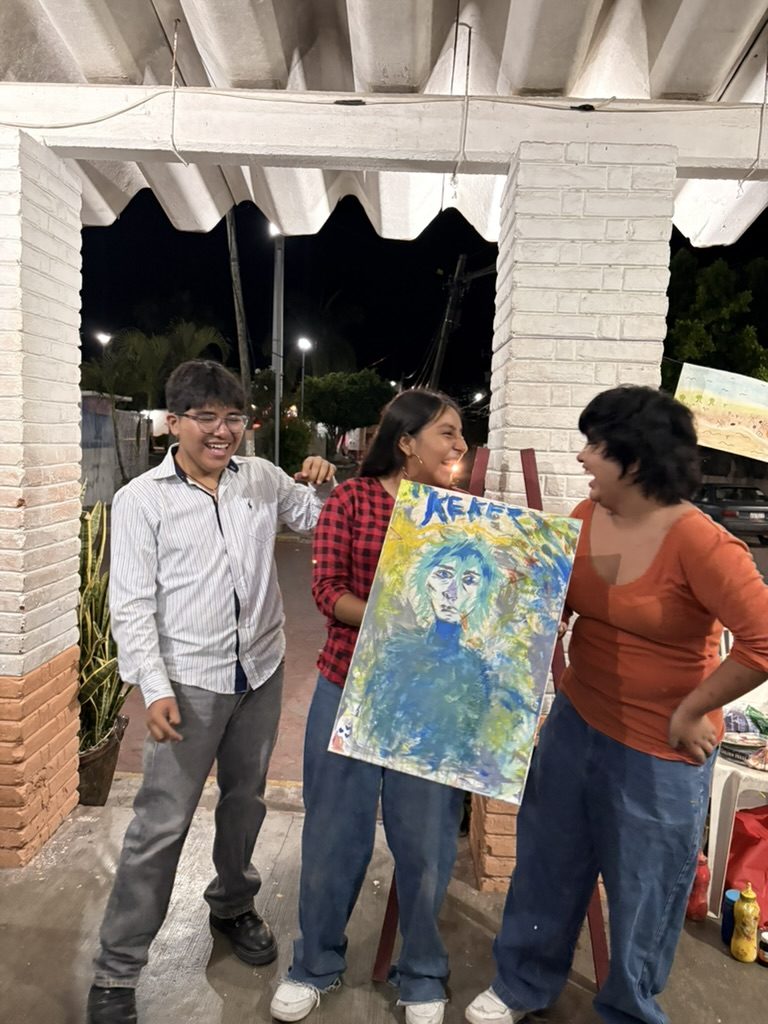 Tres jóvenes se ríen bajo el techo de lámina; una sostiene un retrato pintado en azules
