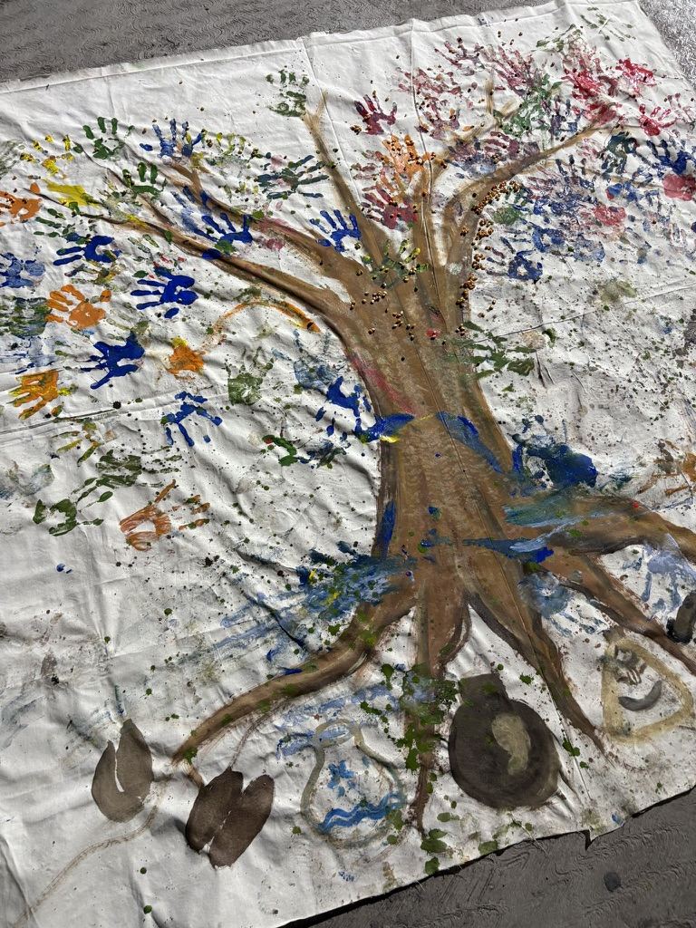 Una manta blanca extendida en el suelo con un &aacute;rbol pintado; el follaje y las ra&iacute;ces est&aacute;n hechos de huellas de manos de muchos colores