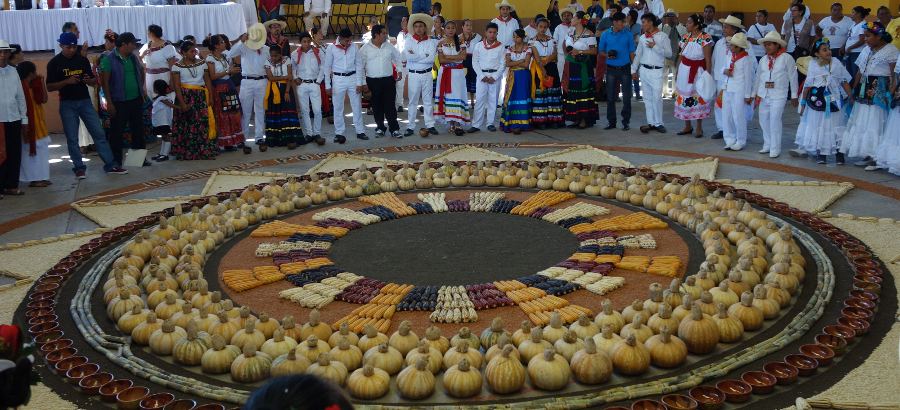 Gente de pie alrededor de un c&iacute;rculo grande hecho en el suelo con ma&iacute;z de varios colores, calabazas, semillas y cazuelas de barro, dentro de un auditorio