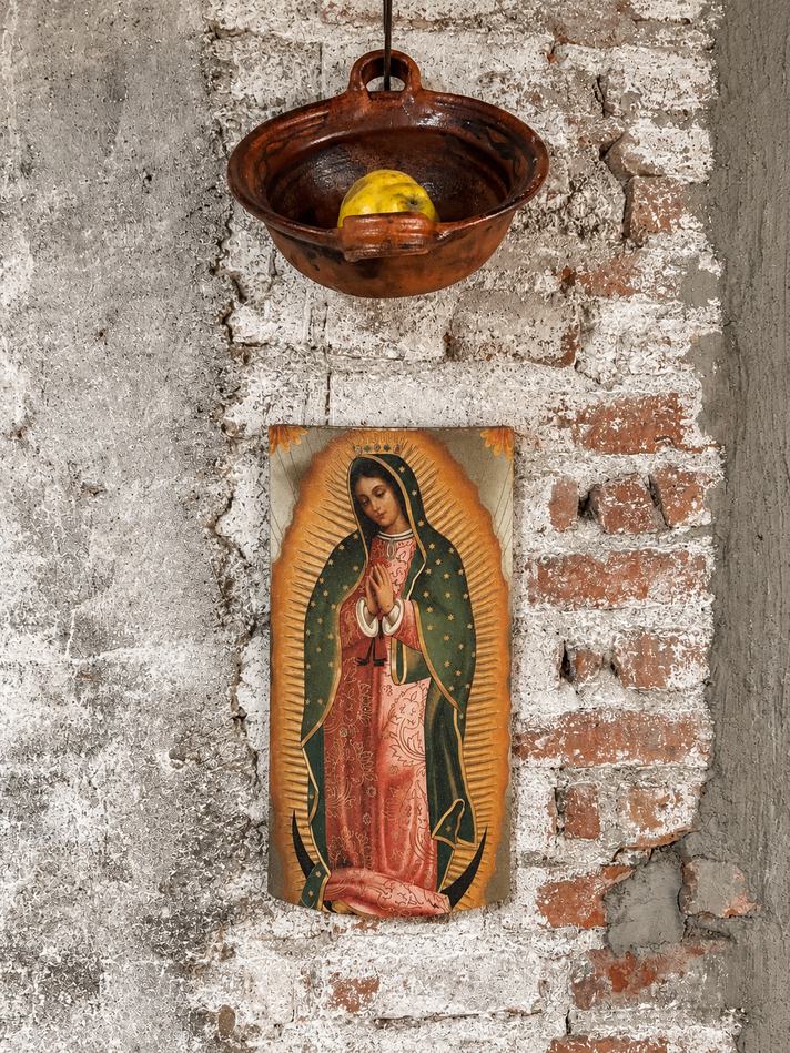 Un cuenco de barro colgado con un membrillo dentro y, más abajo, una imagen impresa de la Virgen de Guadalupe sobre una pared de ladrillo encalado