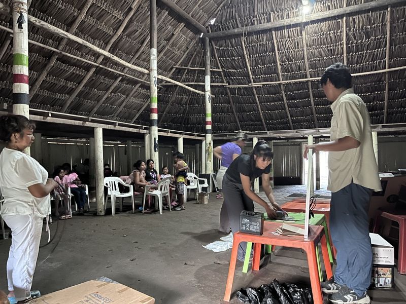 Una mujer sostiene un cable mientras, al fondo, otra conecta una batería sobre una mesa y un joven sujeta un panel de pie