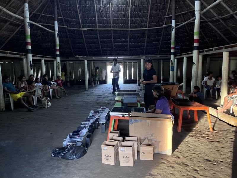 Dentro de la maloca, los componentes de los doce kits en fila sobre el suelo: baterías, reguladores y cajas, con un panel apoyado en una mesa y familias sentadas alrededor