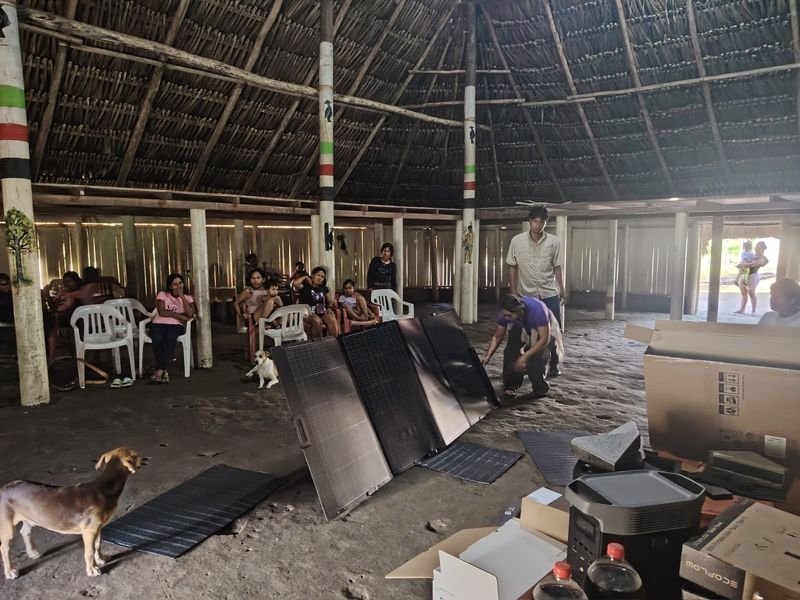 Tres paneles desplegados sobre el suelo de la maloca junto a una estación portátil en su caja; un perro pasa delante y la gente mira desde las sillas