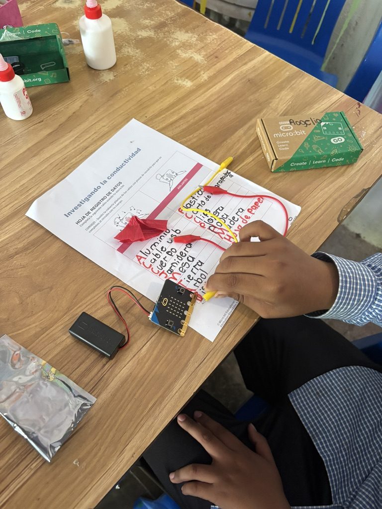 Una mano escribiendo en una hoja de registro sobre la mesa, con una micro:bit, un portapilas y una caja de micro:bit al lado
