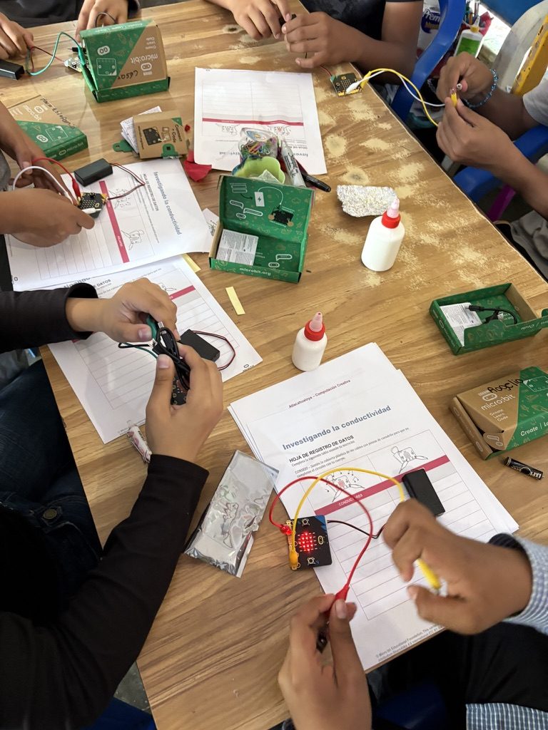Varias manos alrededor de una mesa con micro:bits, portapilas, cables de caim&aacute;n, cajas de micro:bit y hojas de registro repartidas
