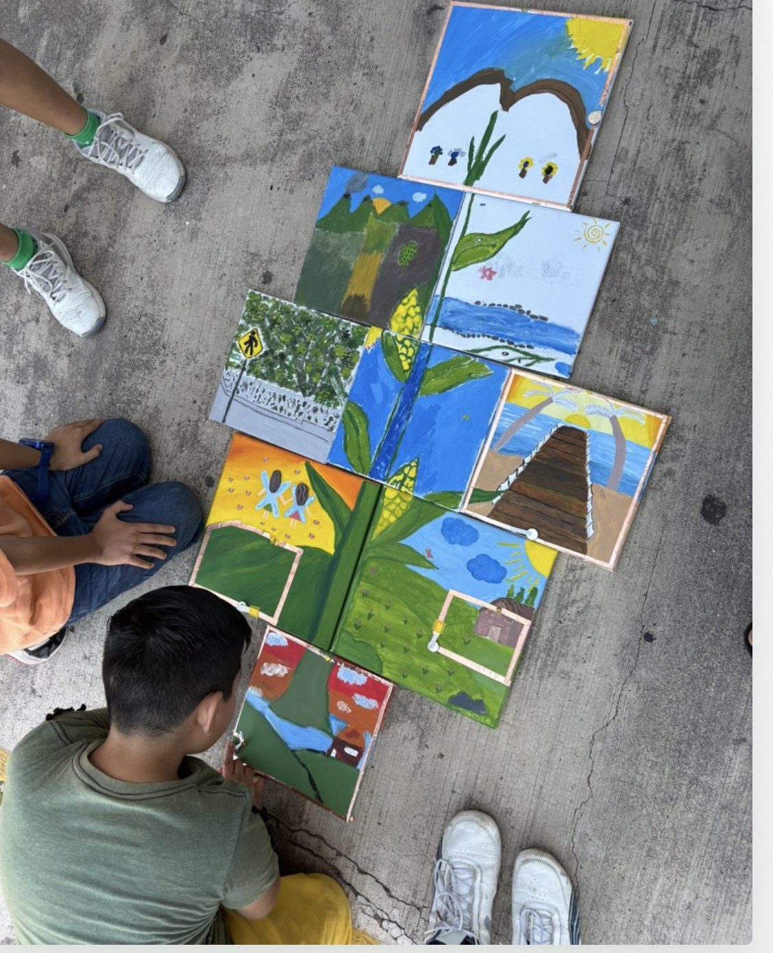 Lienzos pintados puestos sobre el concreto formando un solo paisaje continuo: hileras de ma&iacute;z, cerros verdes, una laguna azul, una pir&aacute;mide escalonada, un sol y nubes. Dos j&oacute;venes est&aacute;n en cuclillas en las orillas, todav&iacute;a acomodando las piezas