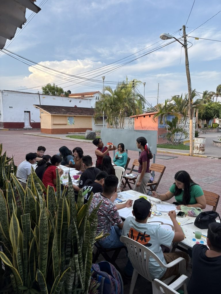Mesas puestas en la calle al atardecer, un grupo de j&oacute;venes pintando, con plantas en primer plano y casas atr&aacute;s