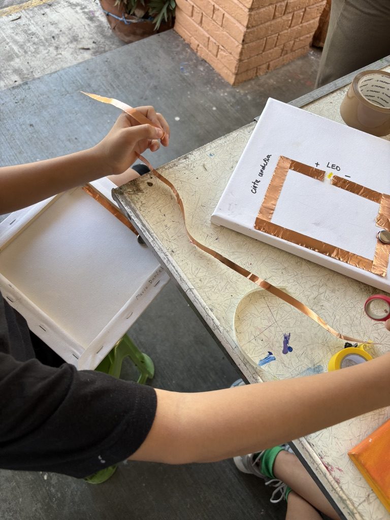 Una mano pintando un lienzo blanco que tiene cinta de cobre pegada alrededor de la orilla, con el nombre Leo escrito en una esquina; cinta, pinceles y un rollo sobre la mesa