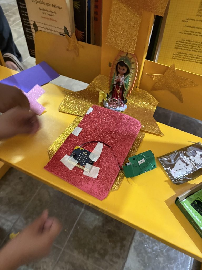Una micro:bit pegada con cinta sobre un tapete rojo con brillos en una mesa amarilla, junto a una figura peque&ntilde;a de la Virgen en un nicho dorado, con un portapilas verde y papel aluminio al lado