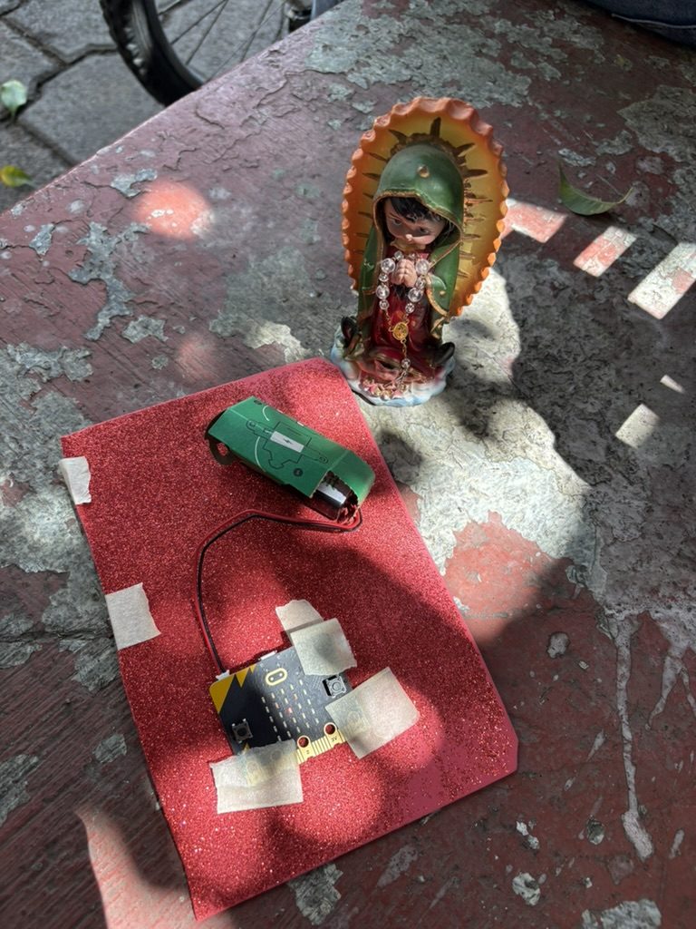 El mismo tipo de dispositivo puesto afuera sobre piedra, pegado a un tapete rojo junto a una figura de la Virgen de Guadalupe, con luz de sol entre las sombras