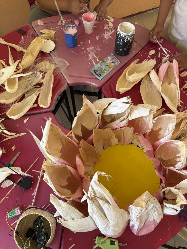 Una mesa cubierta de p&eacute;talos de totomoxtle acomodados en una flor grande con un disco amarillo al centro; alrededor hay botes de pintura, una placa peque&ntilde;a y cables