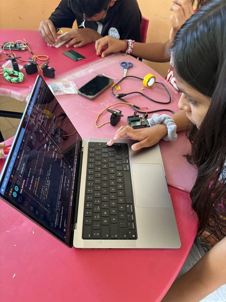 Una joven frente a una laptop con una micro:bit, un servo y caimanes sobre una mesa rosa