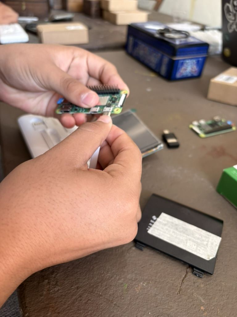 Dos manos sosteniendo una Raspberry Pi Zero sobre la mesa, con la pantalla y la bater&iacute;a al lado