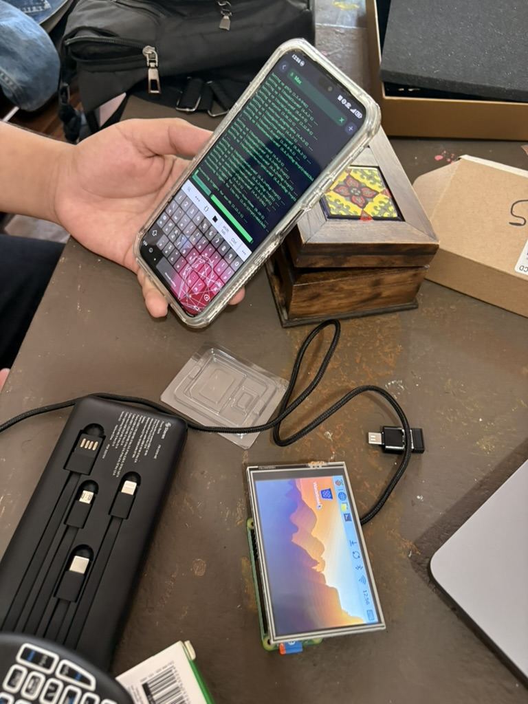 Una mano sosteniendo un tel&eacute;fono con una terminal abierta, junto a una de las computadoras encendida sobre la mesa
