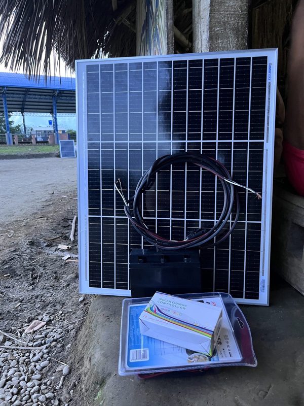 Un panel solar apoyado sobre una caja en el suelo en Maguaré, con el río y un techo de palma detrás