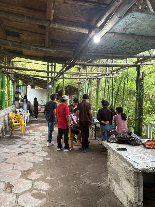 Un grupo de pie bajo un techo de madera y bambú, mirando algo sobre una mesa, con una lámpara encendida
