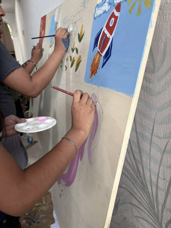 Dos jóvenes pintan un mural sobre madera: un cohete, mazorcas y un cielo azul